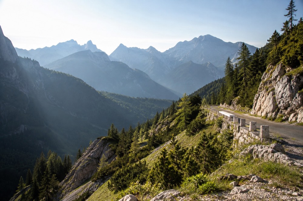 A mountain road curving along a high green ridge with layered blue peaks fading into the distance