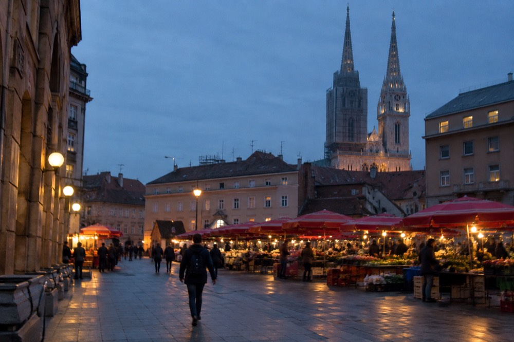 A wide city square with market umbrellas and a cathedral tower behind the rooftops
