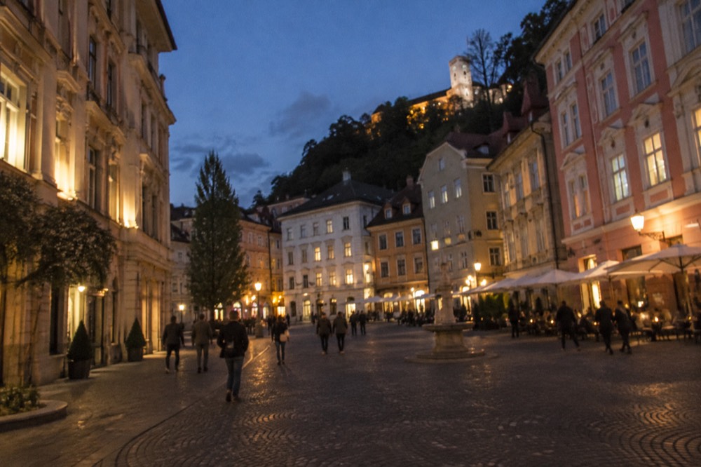An illuminated old town square at dusk with people walking