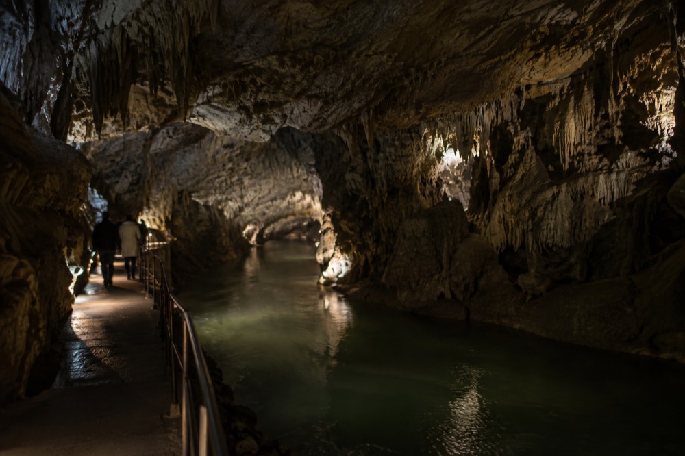 The interior of a karst cave passage lit along a walkway beside an underground river