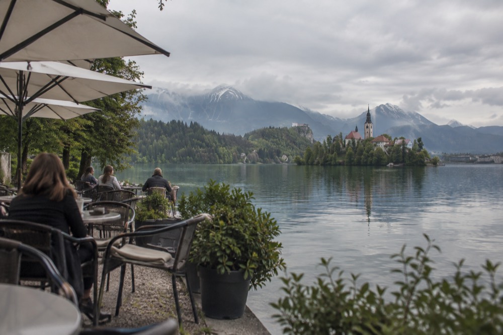 A café terrace with small tables beside a calm lake with a wooded island in the distance
