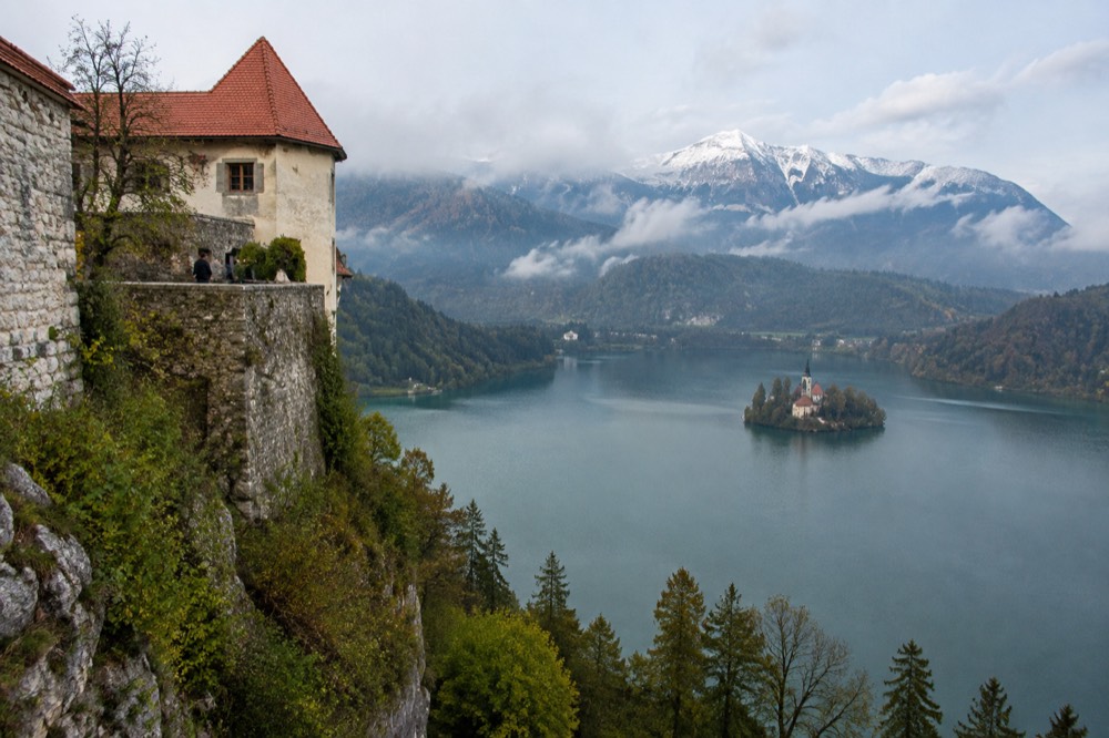 A cliff-top castle above a lake with a small island church and mountains in the distance