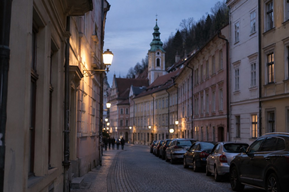 A street of parked cars on the edge of Ljubljana's car-free historic centre, with pastel facades and a church spire behind