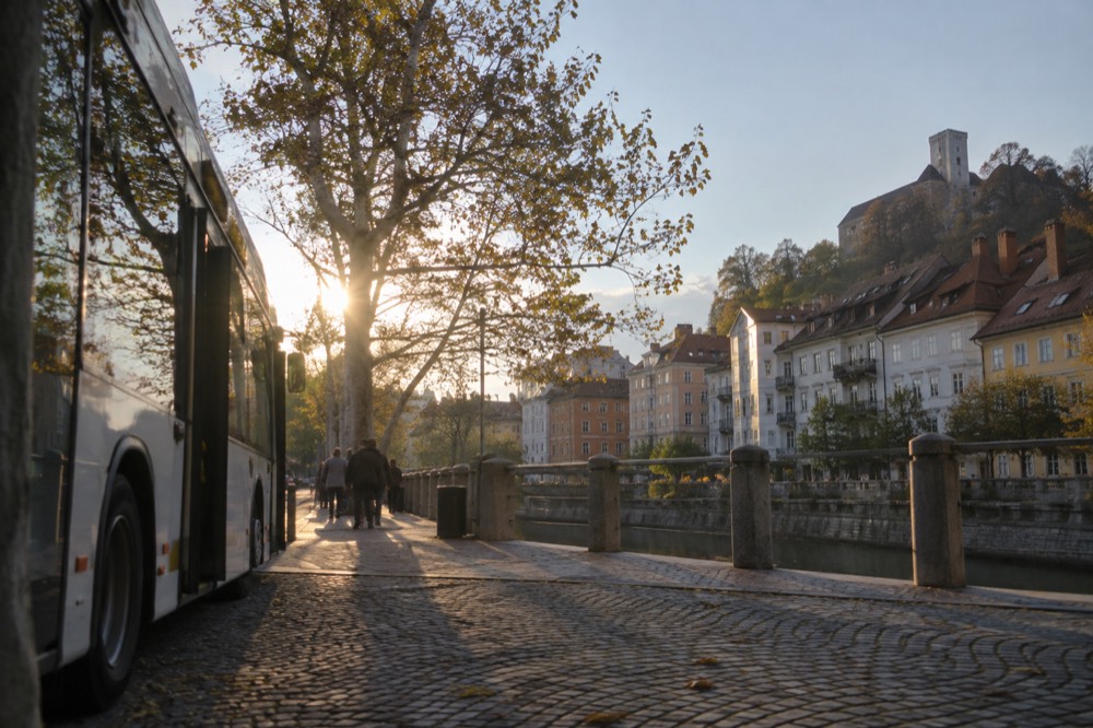 A Ljubljana city bus parked at a stop beside the Ljubljanica river with the old town in the background