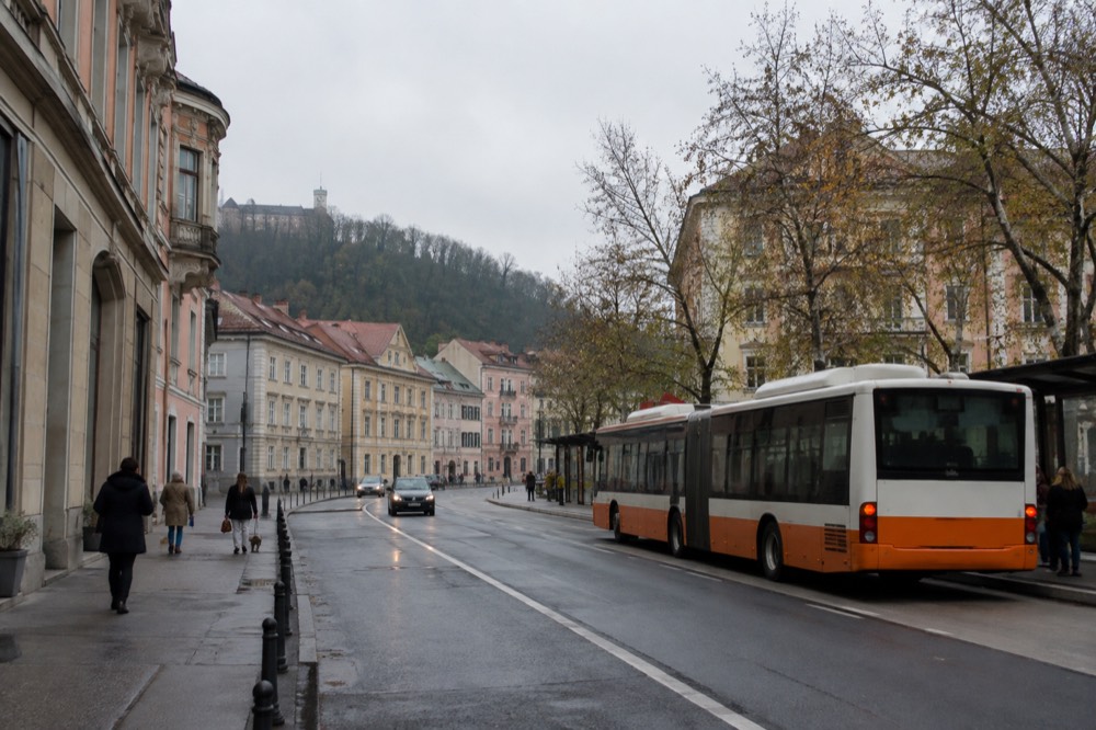 An orange and white city bus at a stop on a wide avenue with apartment buildings behind it