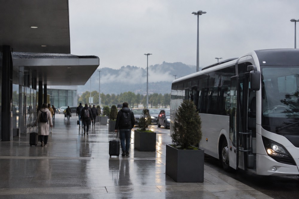 A small airport terminal forecourt with a shuttle bus parked at the kerb and hills behind it.