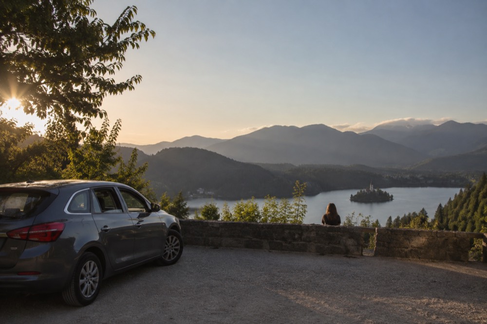 A hire car parked at a scenic overlook with forested mountains and a lake in the distance.