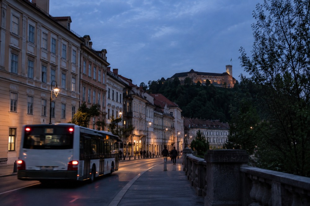 A city bus pulling away from a stop on a wide Ljubljana street with the castle hill behind