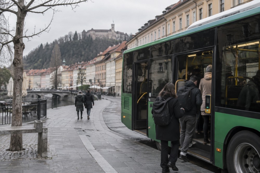 A green Ljubljana city bus at a stop with passengers boarding by a contactless reader