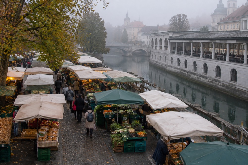 Outdoor food stalls with produce and pastries beside a riverside market square