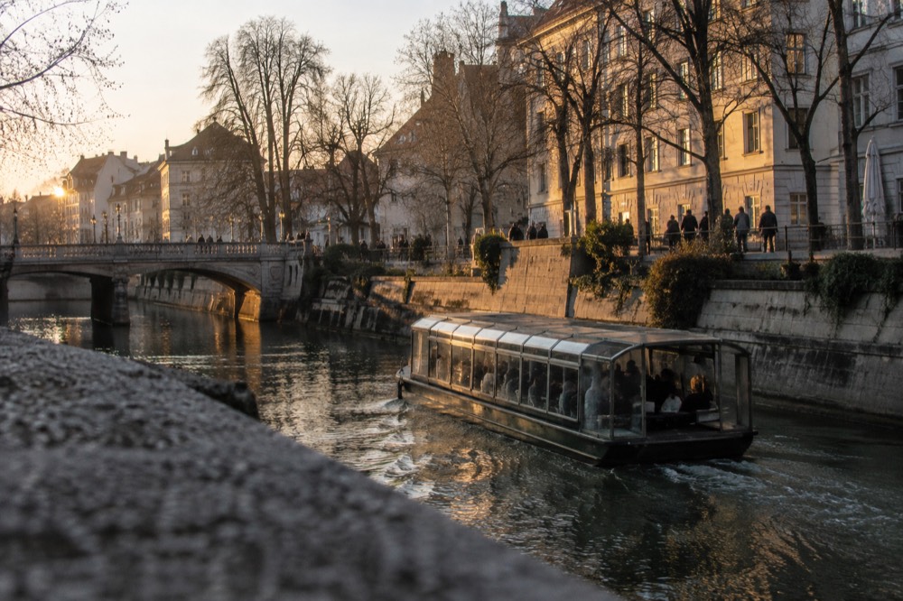 A covered river boat moving past lit riverside buildings on the Ljubljanica
