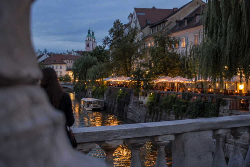 Riverside terrace bars lit by string lights along the Ljubljanica embankment at dusk
