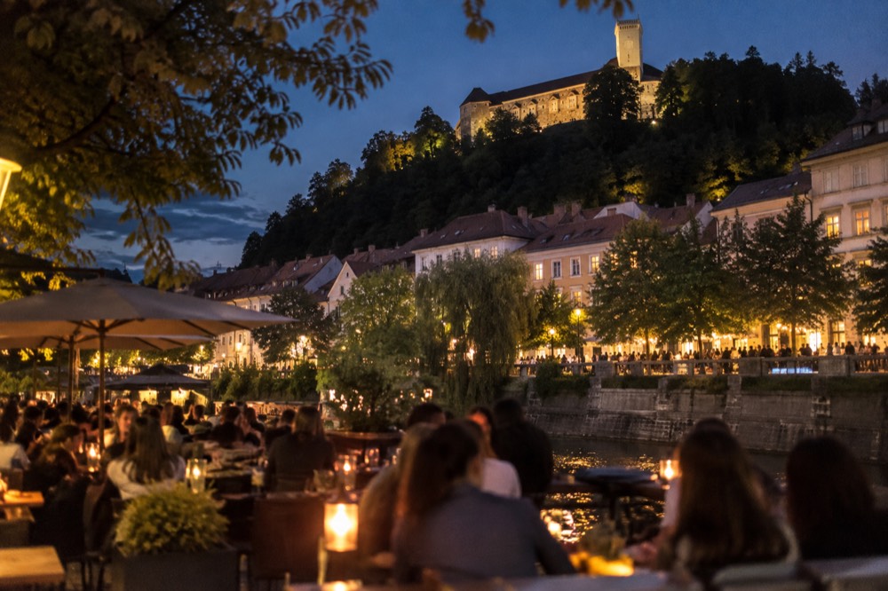 Crowded bar terrace on the Ljubljanica embankment with the Ljubljana Castle hill lit in the background