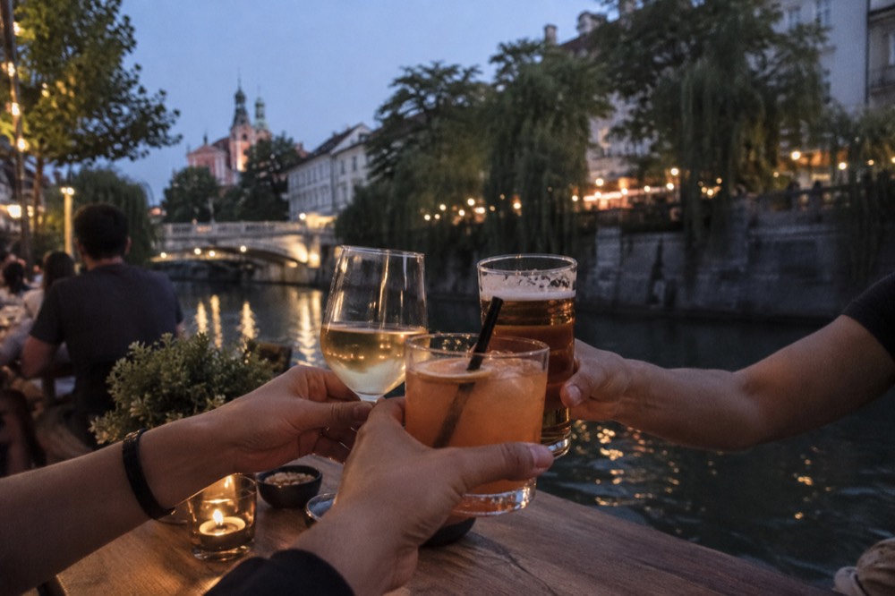 Group of friends toasting glasses at a riverside bar table in the evening