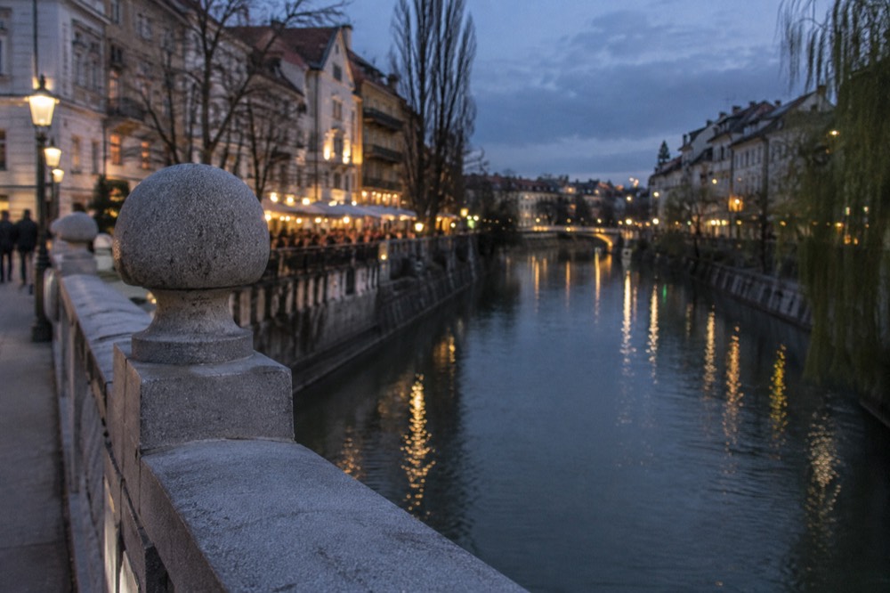A stone bridge over the Ljubljanica river at dusk with lit riverside facades