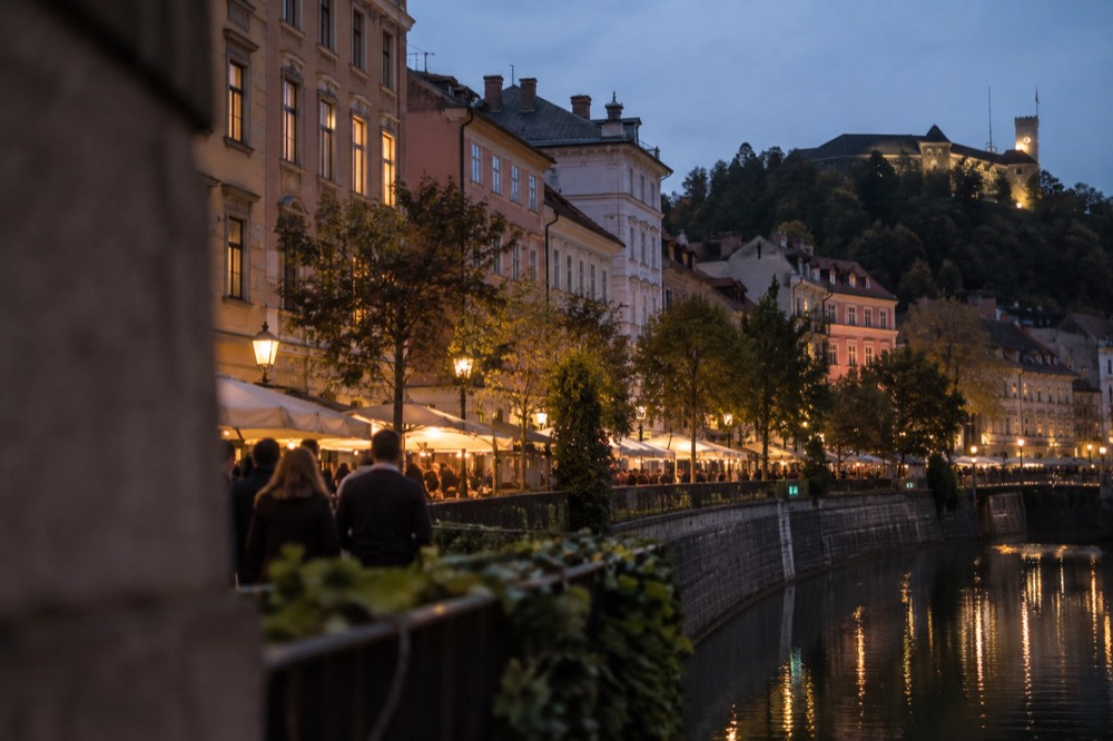 Evening view of a lit market square with people at food stalls beside a river