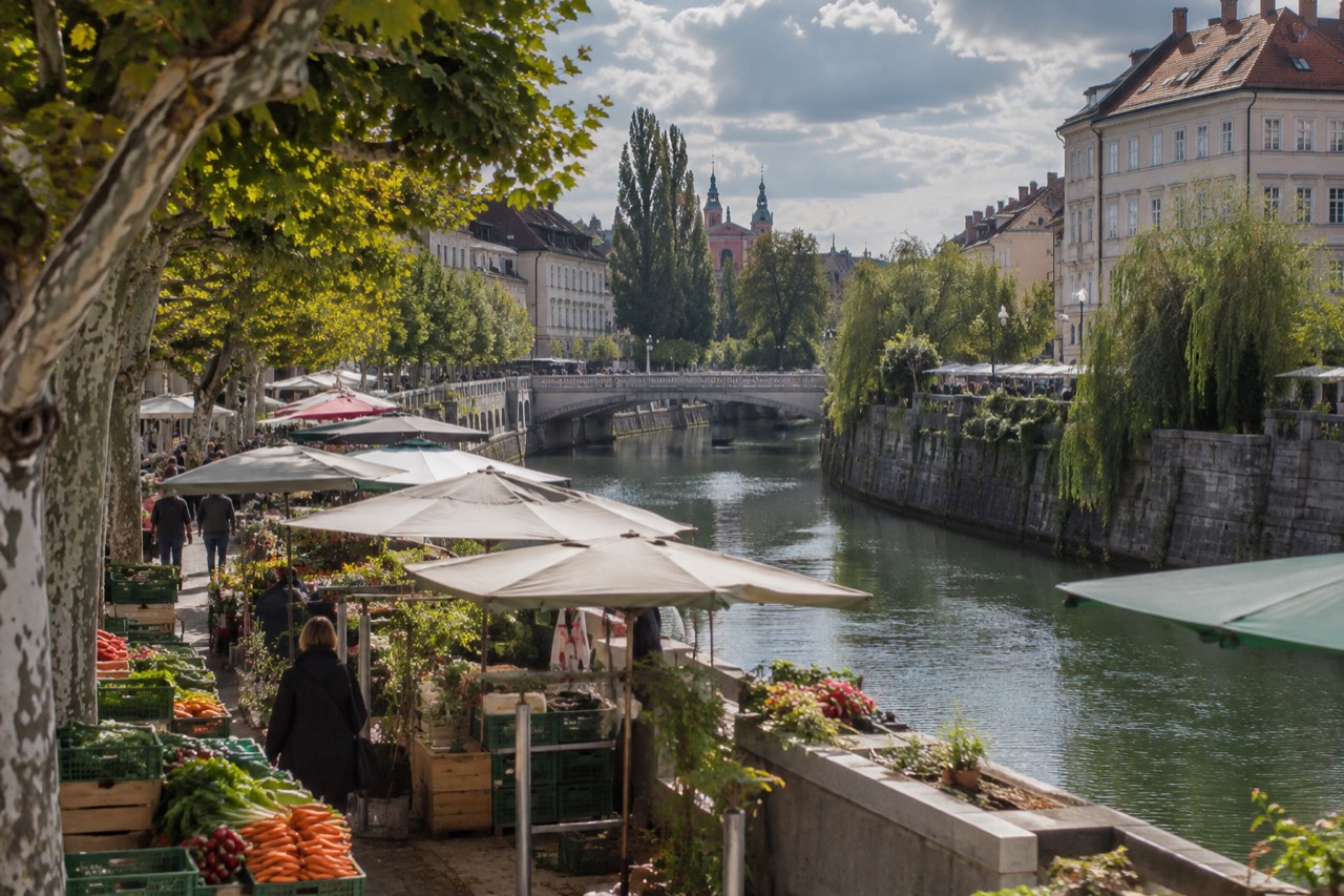 Market stalls with fresh produce beside a river in a pedestrian city centre on a bright morning