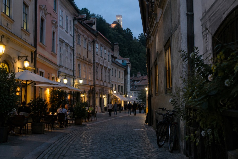 A narrow cobbled street in Ljubljana with restaurant tables and lanterns in the evening